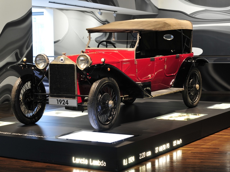 A vintage red Lancia Lambda from 1924 with a tan convertible top and black accents is displayed on a pedestal, showcasing its early 20th-century luxury automobile design.