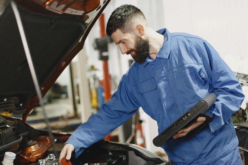 An auto mechanic in blue coveralls inspecting a car's engine in a garage.