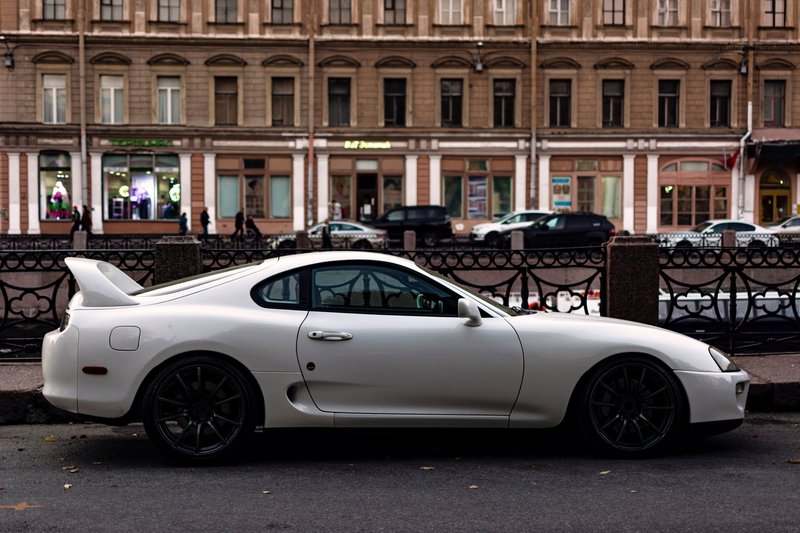 Classic white Toyota Supra parked on a bustling city street, blending retro style with urban architecture.