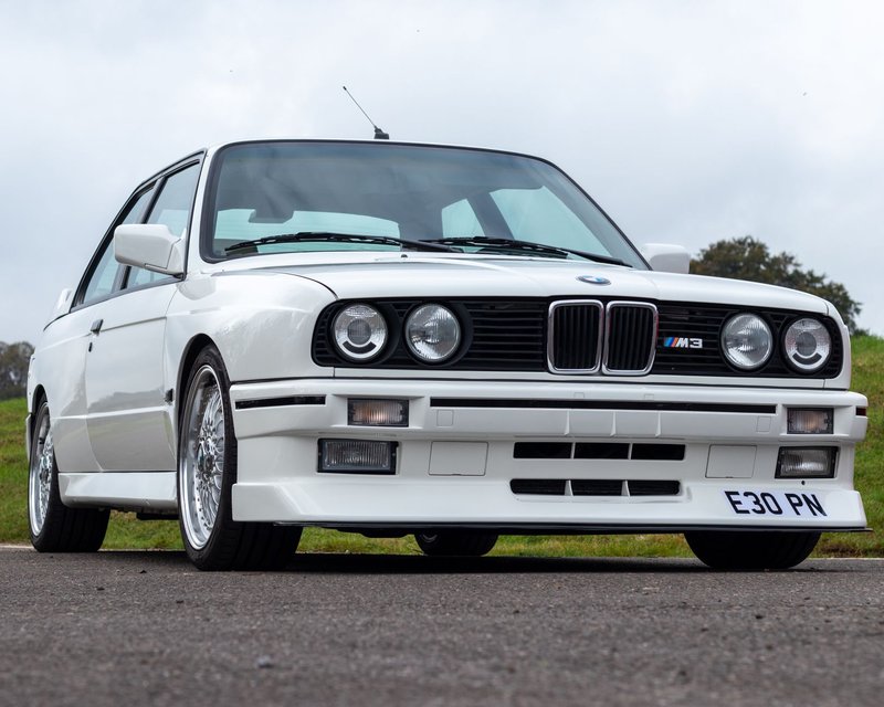 A white classic car with prominent headlights parked on a road in Honiton, England.