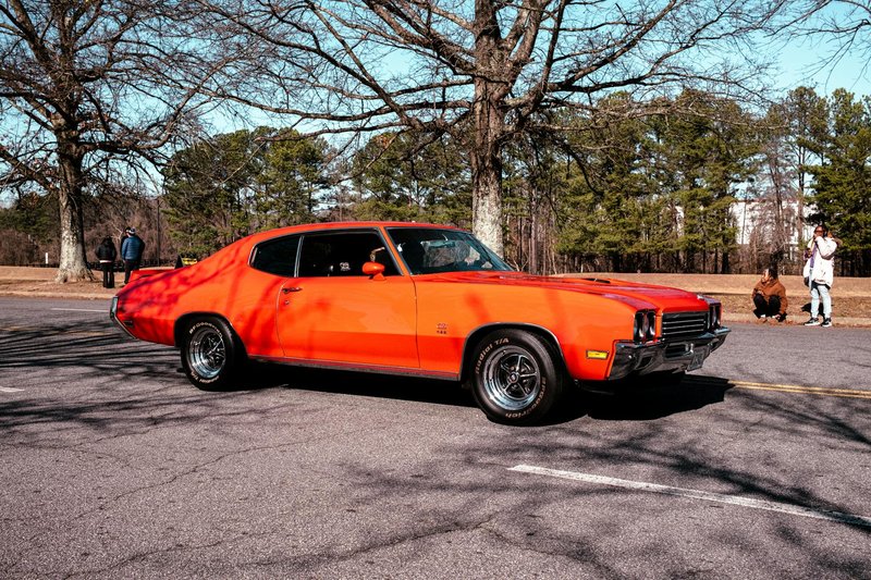 Classic orange muscle car parked amidst trees in a sunlit park setting.