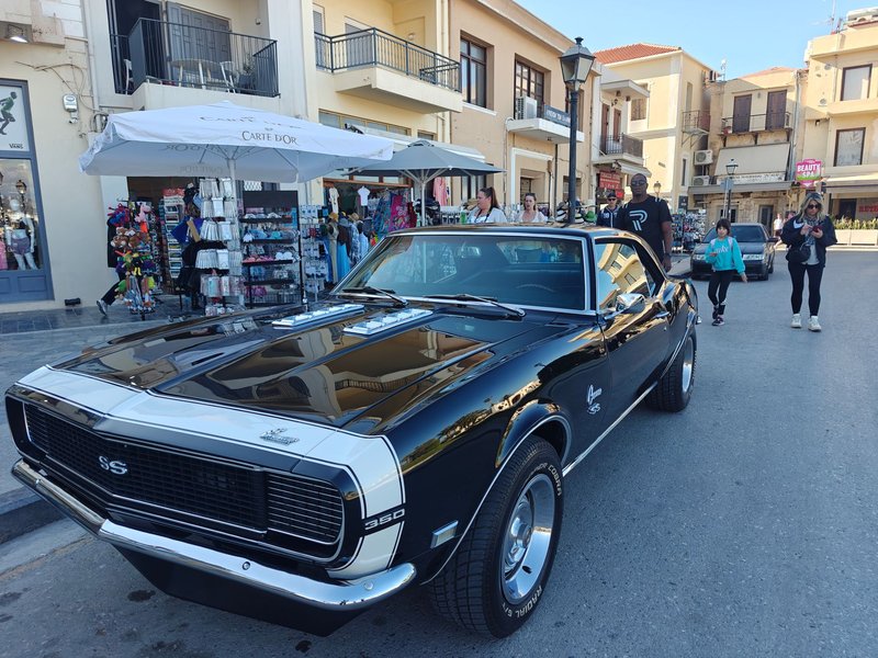 Classic black car parked in a vibrant Grecia street scene with pedestrians.