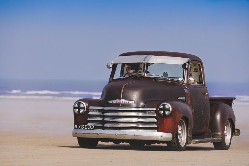 Classic Chevrolet pickup truck driving on a sandy beach with ocean view.