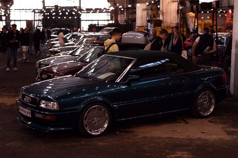 A classic Audi cabriolet showcased at an indoor car exhibition with a bustling crowd.
