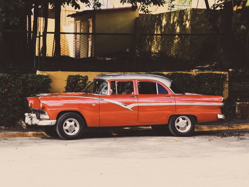 Classic red Ford Customline sedan parked beside a sidewalk with lush greenery.
