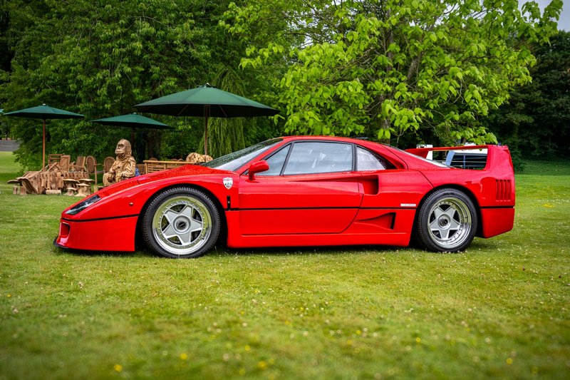Classic red Ferrari F40 parked in a lush green park with wooden sculptures.