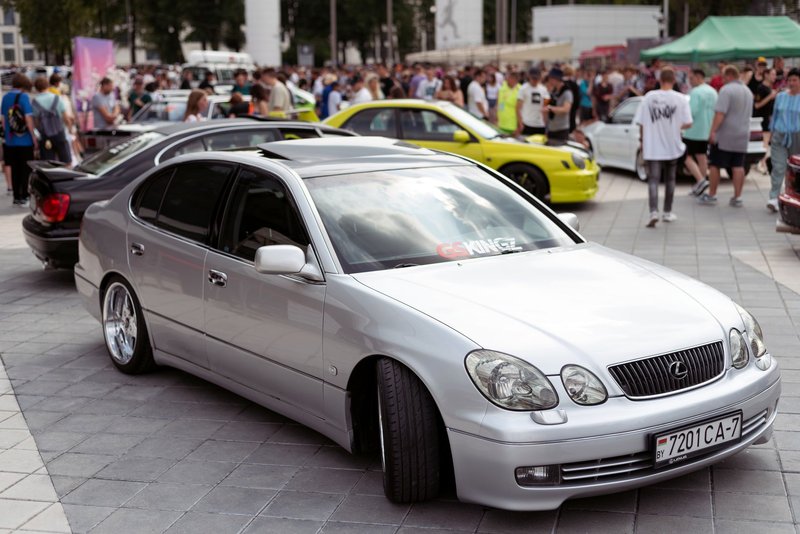 A silver Lexus GS showcased at an urban car show with a crowd in the background.