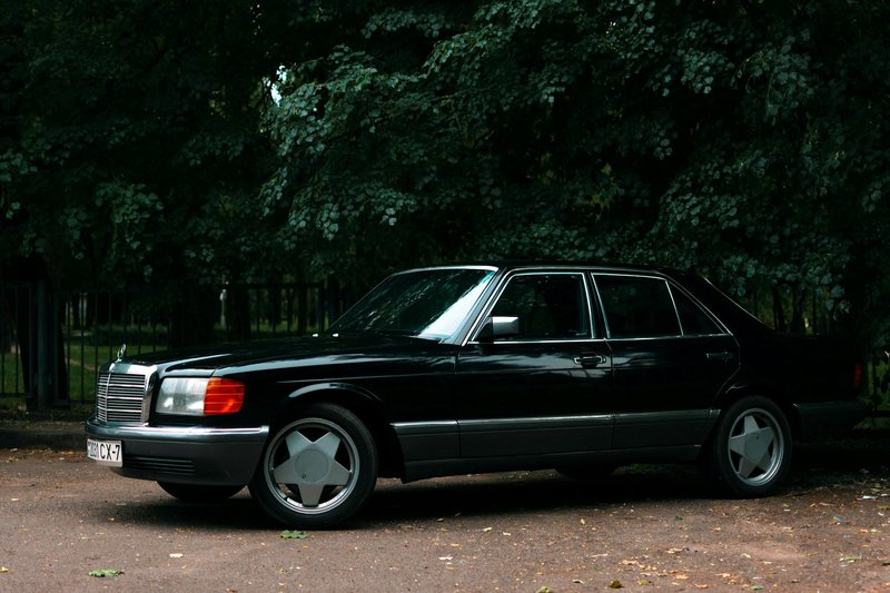 A vintage black Mercedes-Benz W126 car parked in a shaded outdoor setting.