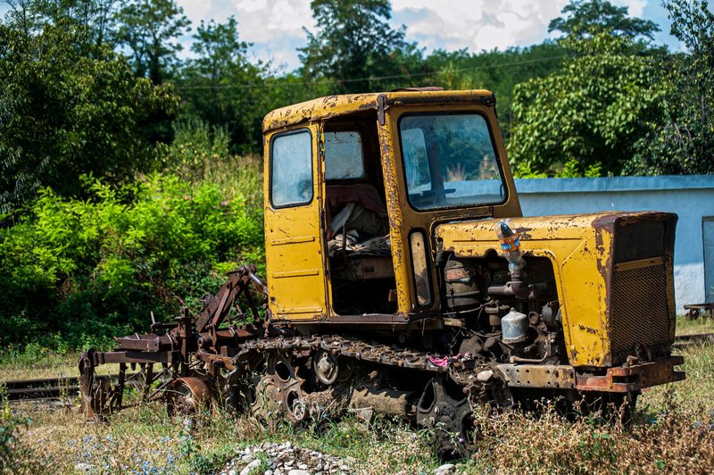 An old, rusty yellow tractor sits abandoned outdoors surrounded by greenery.