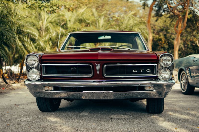 Front view of a classic red Pontiac GTO parked in a sunny park with trees in the background.