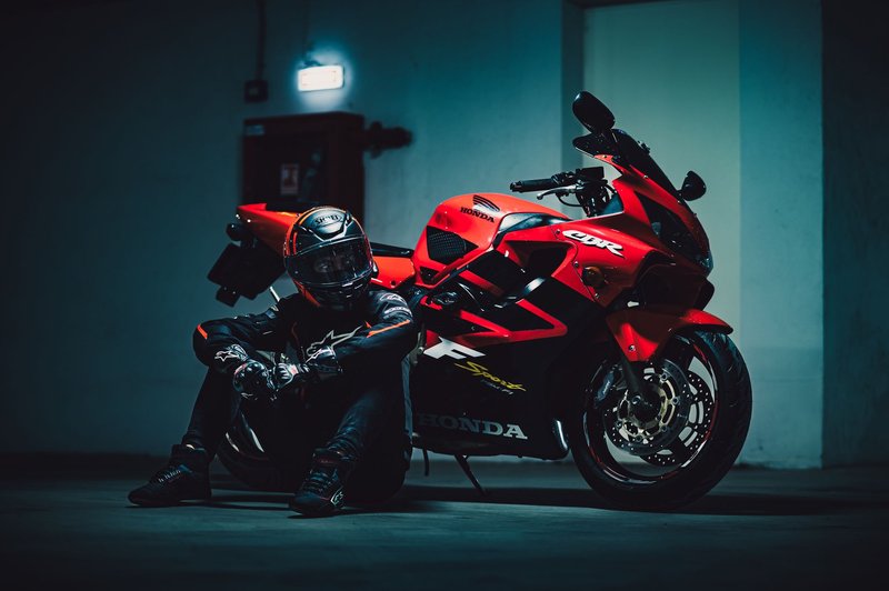 Motorcyclist seated next to a red sport bike in dimly lit garage.