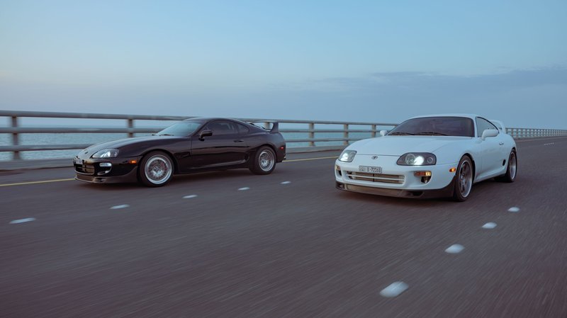 Black and white Toyota Supra cars racing on a coastal road during the day.
