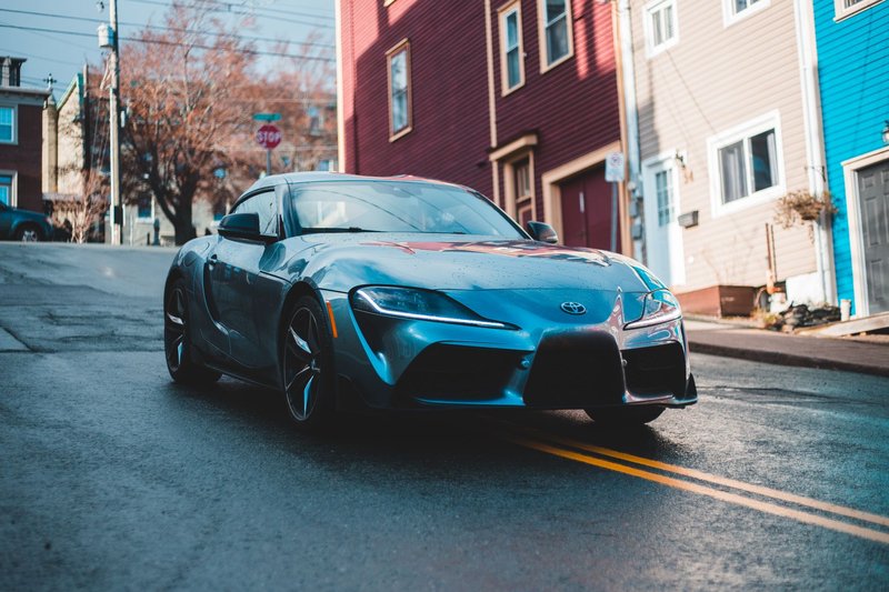 A blue Toyota Supra parked on a wet city street, showcasing its elegant design against colorful houses.