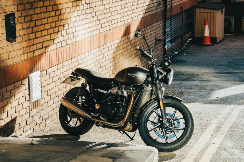 A classic Triumph motorcycle parked under sunlight in an urban alleyway with a brick wall background.