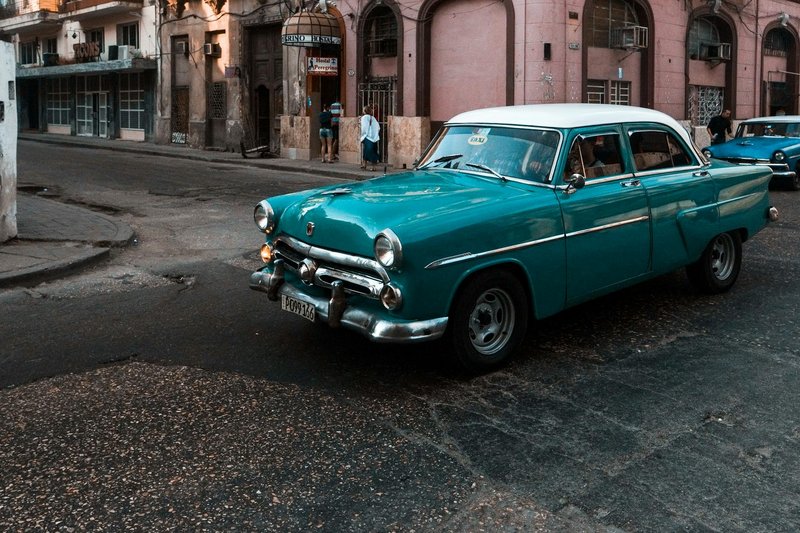 Classic blue car driving on a historic city street with buildings in the background.