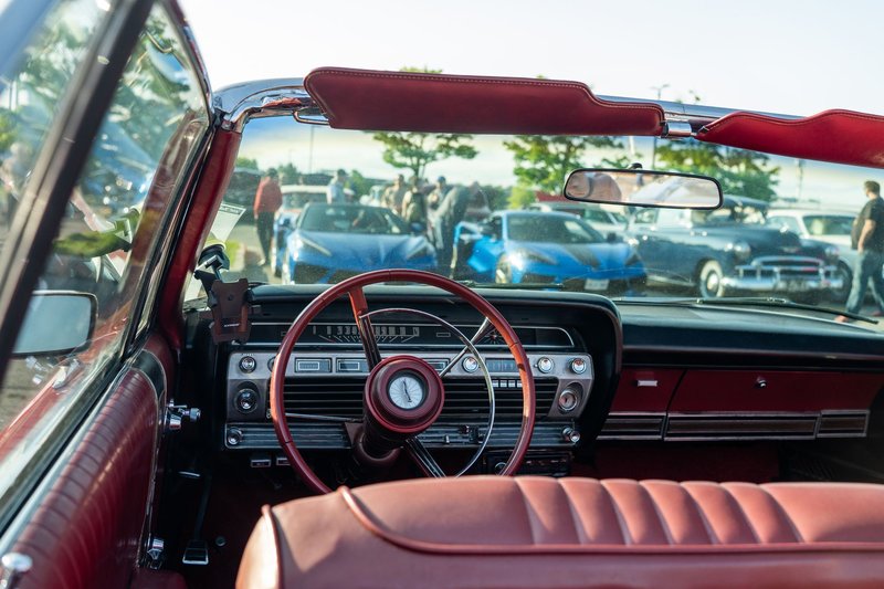 A vintage car interior view at an outdoor auto show featuring classic cars in the background.