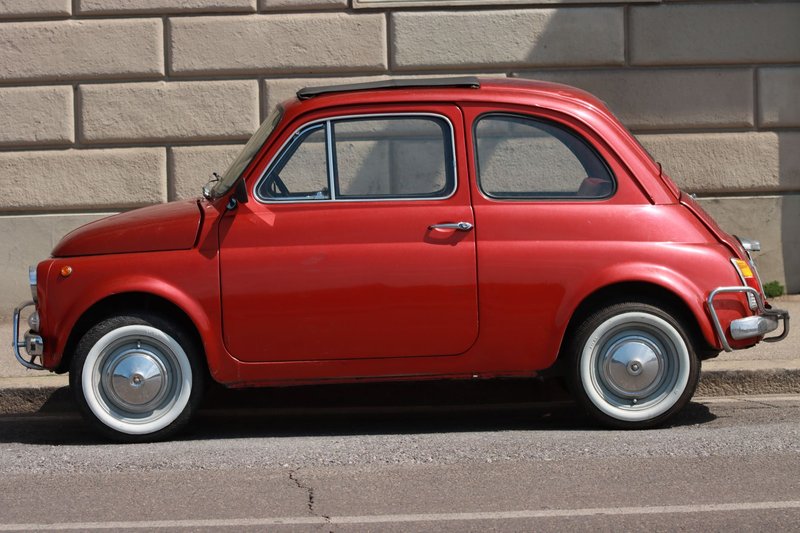 Side view of a classic red Fiat car parked against a brick wall in an urban setting.