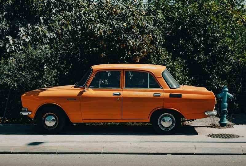 A classic orange Moskvitch 2140 sedan parked on a city sidewalk with lush greenery.