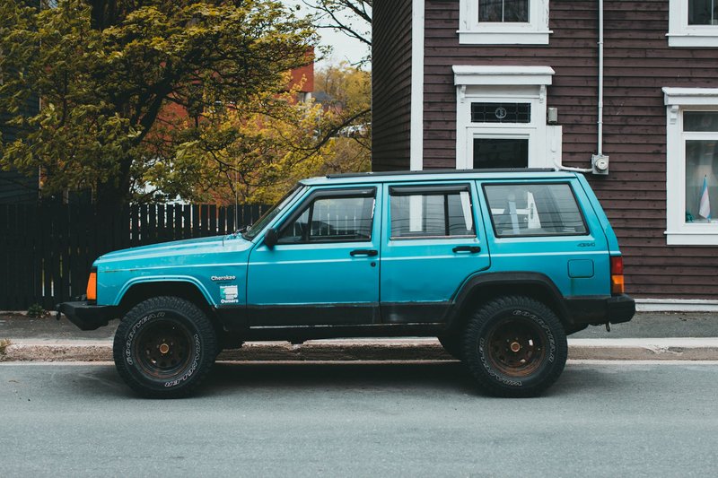 Bright blue vintage Jeep Cherokee parked by a rustic building on a tree-lined street.