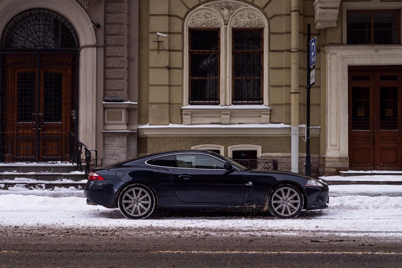 Elegant Jaguar XK parked on a snowy urban street beside a classic building, showcasing winter city scenery.