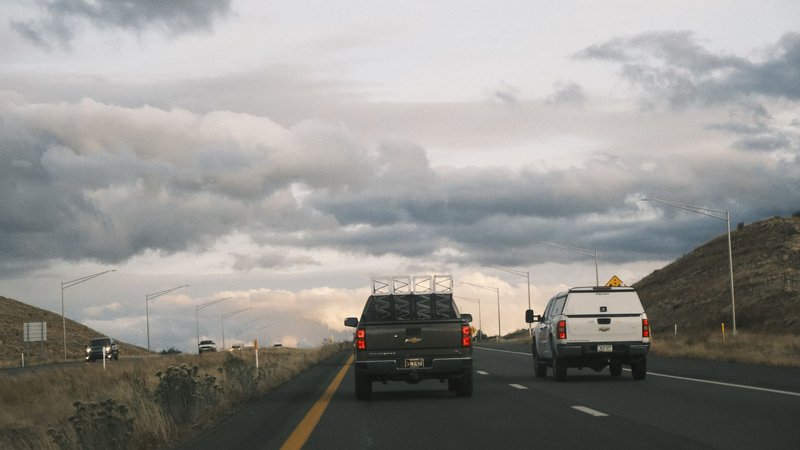 Two pickup trucks drive along a scenic highway under cloudy skies.