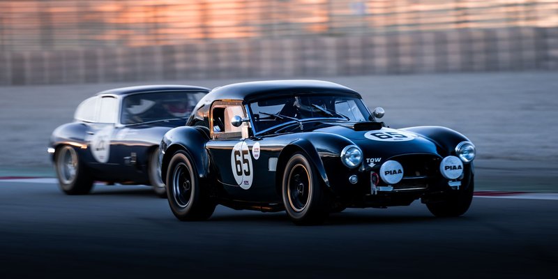 Two classic cars racing intensely on a track during sunset, highlighting speed and competition.