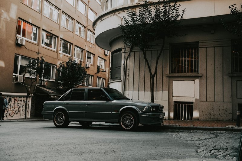 A classic car is parked on a city street, surrounded by urban architecture and trees.