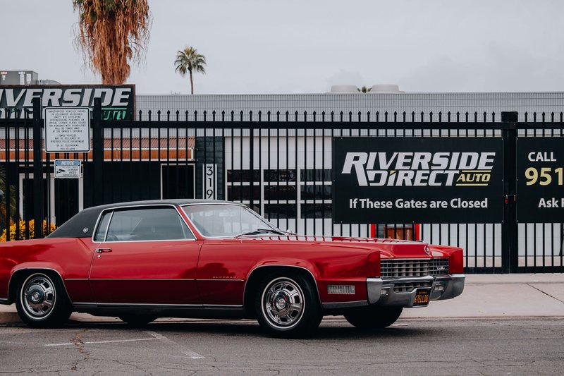 Classic red vintage car parked in front of Riverside Direct Auto in a city setting, showcasing urban automotive style.