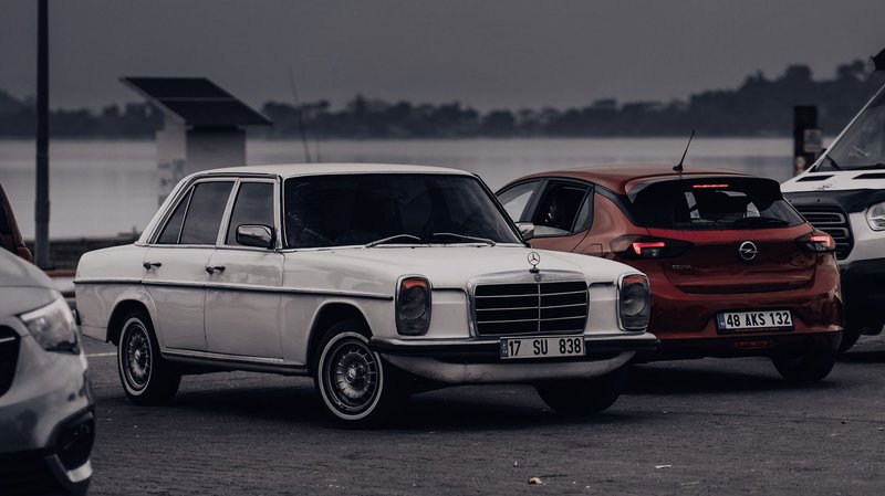 Classic white car parked by a serene lakeside, with other vehicles nearby on a cloudy day.