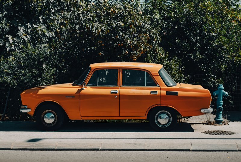 A classic orange Moskvitch 2140 sedan parked on a city sidewalk with lush greenery.