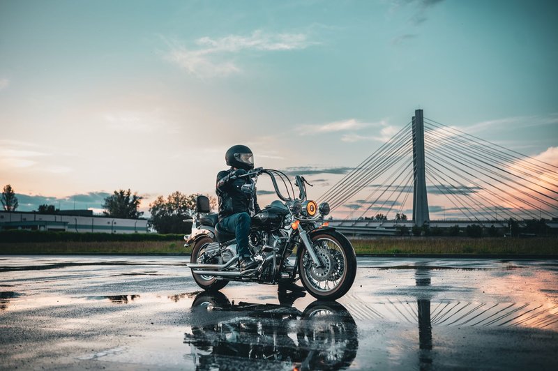 Motorcycle rider in leather gear on a wet road with a bridge in the background during sunset.