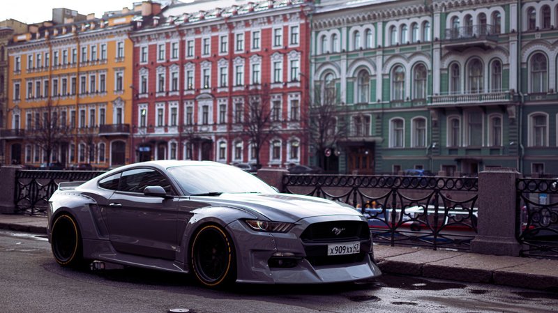A sleek grey sports car parked on an urban street with colorful historic buildings in the background.