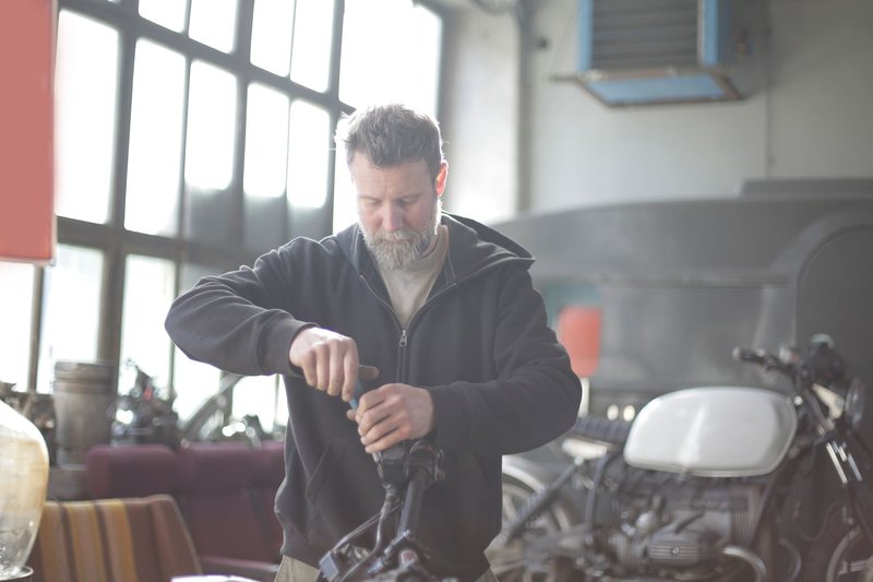 Serious male mechanic in casual wear with gray beard standing in garage and fixing motorcycle steering wheel in back lit