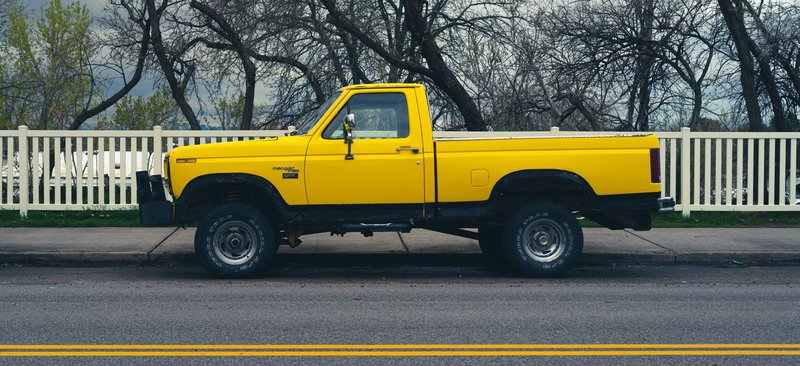 Side view of a parked yellow pickup truck on a city street with trees and a fence in the background.