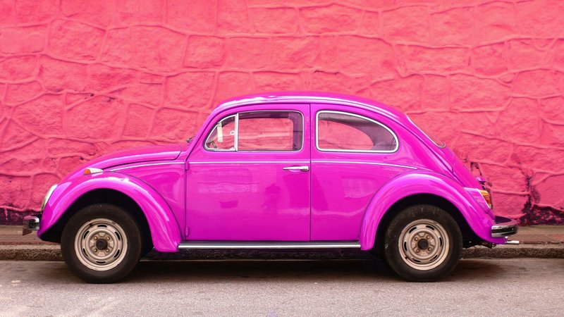 Side view of a vibrant pink classic Volkswagen Beetle parked in front of a pink wall in an urban setting.