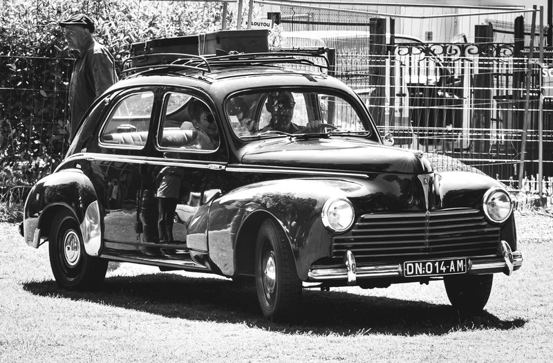 Black and white photo of a vintage Peugeot car with whitewall tires, captured in an urban setting.