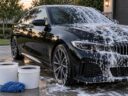 Black car half covered in car wash foam next to two wash buckets and a microfiber mitt