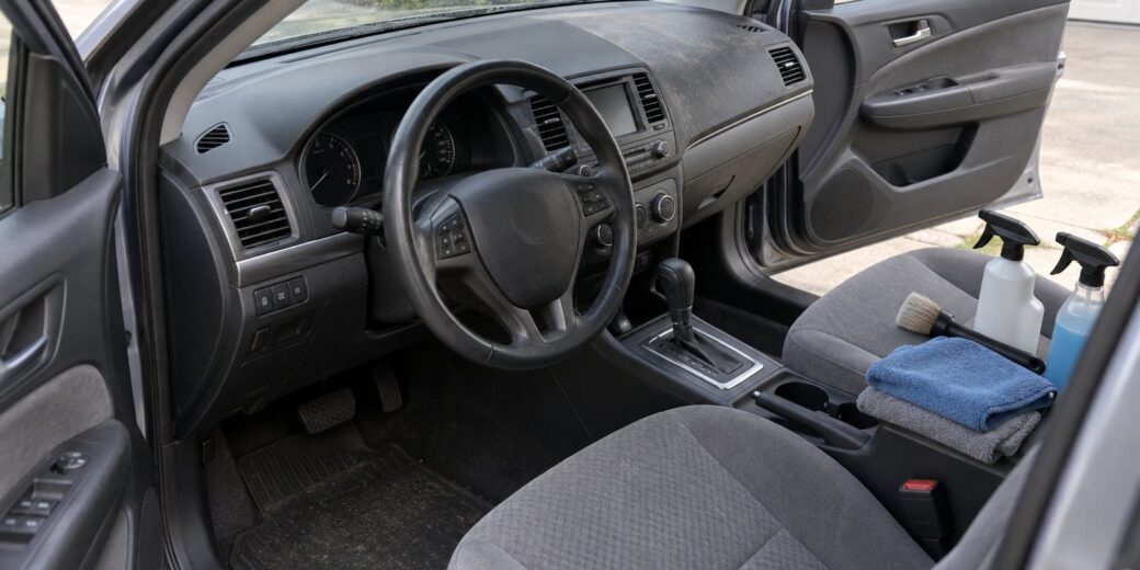 Car interior with cloth seats, unlabeled cleaning sprays, a detailing brush and microfiber towels on the passenger seat