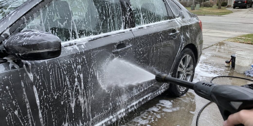 Dark gray sedan covered in soap suds while a pressure washer sprays the door in a driveway