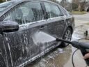 Dark gray sedan covered in soap suds while a pressure washer sprays the door in a driveway