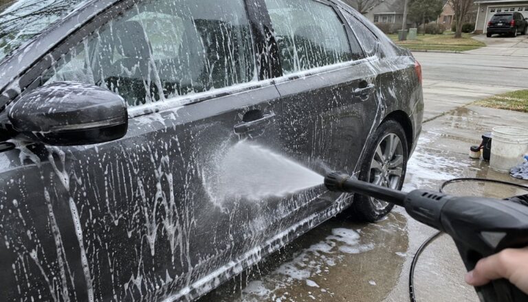 Dark gray sedan covered in soap suds while a pressure washer sprays the door in a driveway