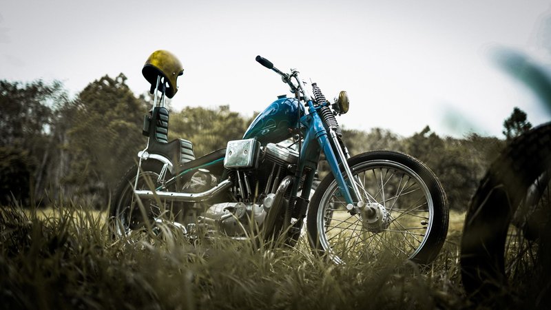 A classic chopper motorcycle parked on a grassy field with a helmet on the handlebars, under a cloudy sky.