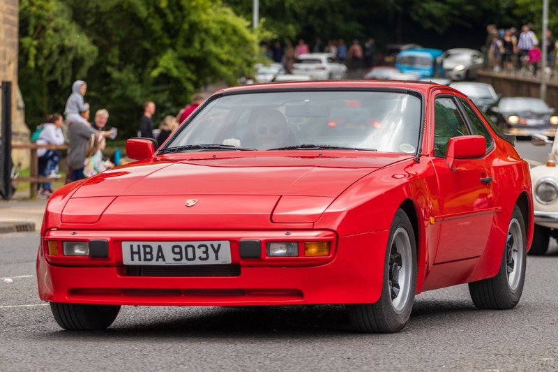 Vintage red Porsche 944 cruising through Morpeth, England, showcasing classic car enthusiasts and street scene.