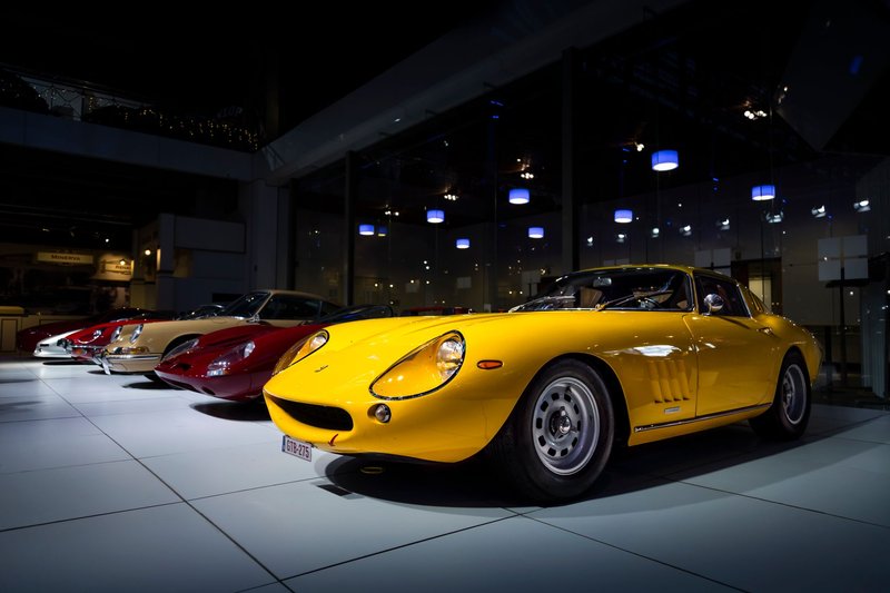 A lineup of vintage sports cars on display indoors, highlighting a yellow classic model under soft lighting.