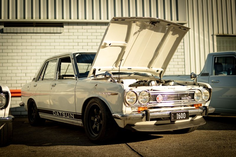 Classic Datsun 1600 with hood open, showcased at a Brisbane car meet under bright sunlight.