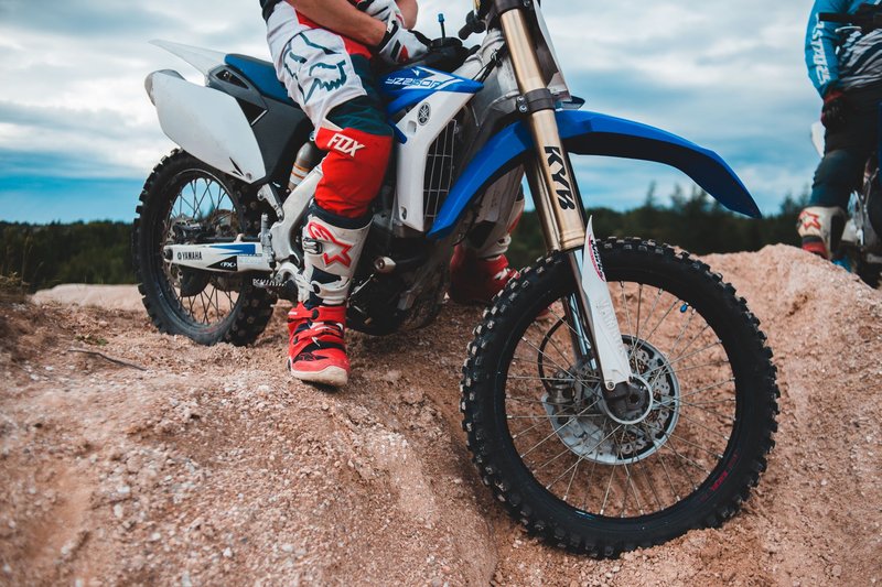 Unrecognizable male racer in sportswear sitting on motorbike on sandy pile against cloudy sky with forest in distance in daylight