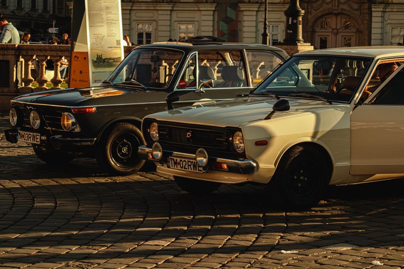 Two vintage cars parked on a cobblestone street in a historic European town during the day.
