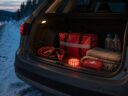 Open car trunk on a snowy roadside at dusk with a red emergency kit bag, jumper cables, LED flare, first aid kit, blanket and water