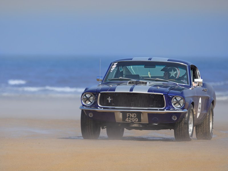 Vintage Ford Mustang GT350 driving on a sandy beach with scenic ocean backdrop.