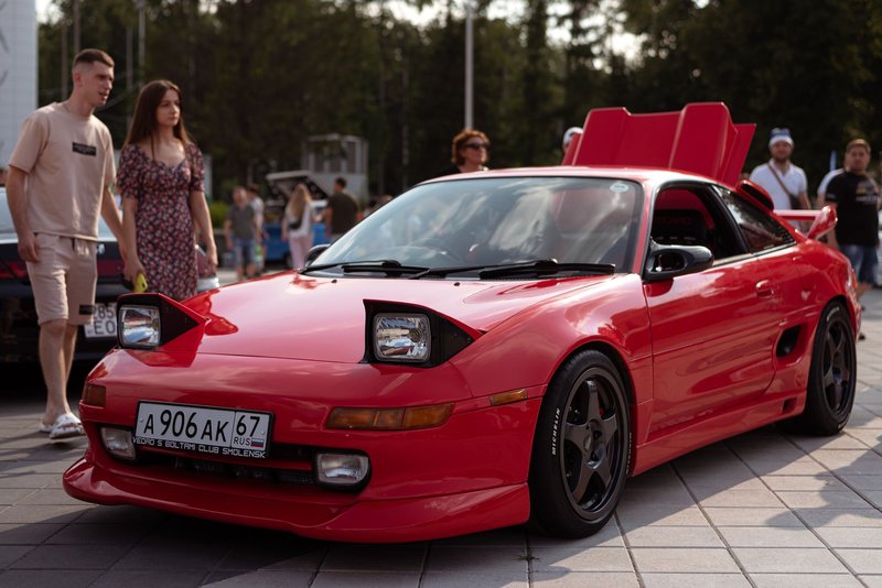 A red Toyota MR2 parked on urban pavement surrounded by people under daylight.